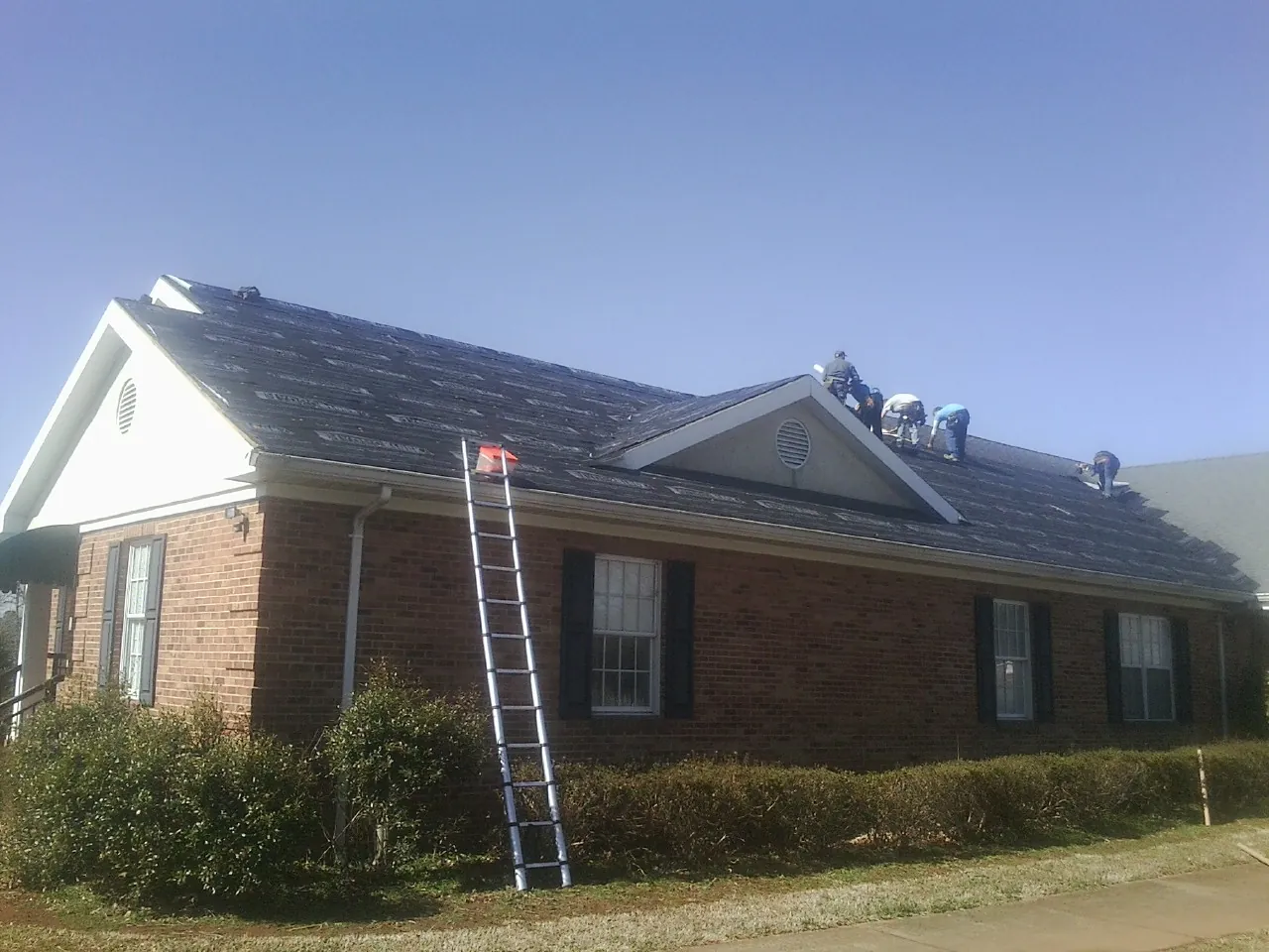 Crew tearing shingles off a brick ranch roof with a ladder set — Metal Roof Repair work by our crew in Marble Hill
