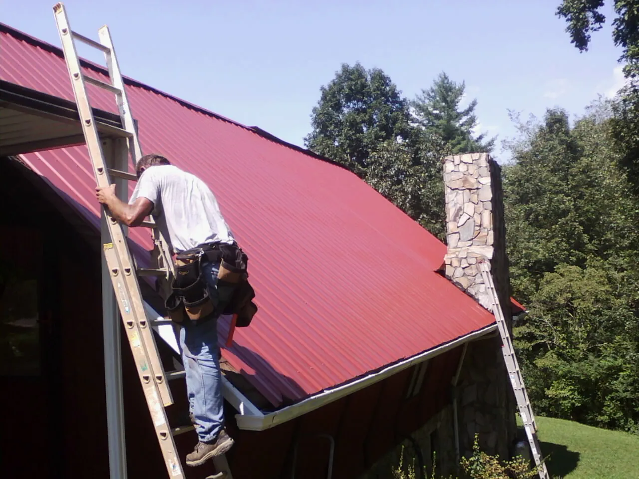 Roofer on a ladder beside a red metal roof and stone chimney — Soffit & Fascia Repair by Roofing Brothers in Marble Hill